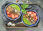 Green smoothie bowls with strawberries, granola, chia and pumpkin seeds, dried figs and nut in wooden tray