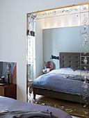 Grey-upholstered bed reflected in mirror with etched ornamental frame