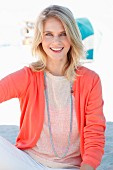 A blonde woman wearing a knitted jumper, a salmon-coloured cardigan and white trousers on the beach