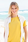 Blonde Frau in gelbem T-Shirt und weißem Pullover über den Schultern am Strand