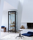 Vase and firewood in black-framed display case next to open fireplace and blue floor cushion on flokati rug