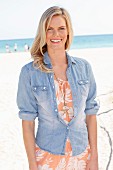 A blonde woman wearing a summer dress with a floral pattern and denim shirt on the beach