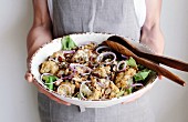 A woman is holding a large bowl of a salad made with roasted cauliflower, lentils, dates, onions, and dates