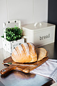 Bread with knife on wooden board, herb pot and bread box in background