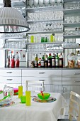 Kitchen counter with mirrored splashback and many glasses on wall-mounted shelves behind colourful place settings on white table