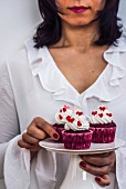 A woman with a white dress holding a cake stand full of red velvet cupcakes