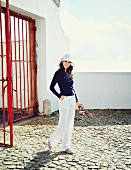 A brunette woman wearing a blue jumper, white linen trousers, a hat and espadrilles