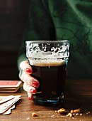 Glass of drunk Guinness with a ladies hand with red nails and a green sweater holding glass at a wooden table surrounded by nuts and playing cards