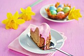 A piece of gugelhupf with a pink sugar glaze, a caramel nest with colourful sugar eggs and daffodils in the background