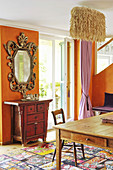 Baroque mirror and Chinese cabinet against orange wall and coconut fibre lamp above dining table