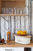 Bread box, vegetables and glasses on shelf against tree-patterned wallpaper
