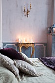 Arrangement of white candles of various shapes on antique gilt console table with scatter cushions on double bed in foreground