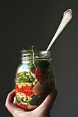 A hand holding a jar of barley salad with balsamic vinegar, roasted tomatoes, baba ghanoush, spinach, cress and almonds
