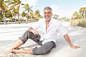 A man with grey hair wearing a white shirt and grey trousers sitting on the beach
