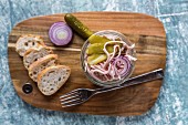 A sausage, red onion and gherkin salad in a glass on a wooden board, with baguette slices