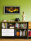 Books on sideboard with white drawers and shelves below framed artwork on light green wall