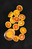 A glass jug of orange juice and halved Moro oranges against a black background