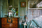 Antique chest of drawers next to metal bed in vintage bedroom with corrugated iron wall