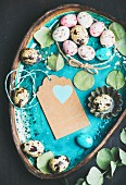 Colorful quail eggs, dried flowers and leaves for Easter holiday over turquoise blue tray with craft paper label