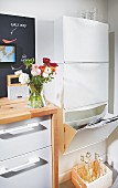 A plastic box for paper and a wooden crate for old bottles in a recycling corner of a kitchen with a white kitchen unit with a wooden countertop and ranunculus flowers in a vase