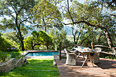Rattan armchairs around set breakfast table on wooden terrace with swimming pool in background
