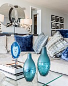 Two blue glass vases and coffee table with glass top in front of sofa with scatter cushions