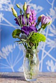 Arrangement of purple flowers in glass vase in front of wall with floral pattern