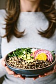 A woman wearing a gray shirt is holding an abundance bowl made up of quinoa, sprouts, lentils, avocados, and chioggia beets