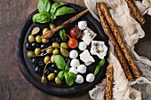 Mediterranean appetizer antipasti board with green black olives, feta cheese, mozzarella, capers, pepper, basil with grissini bread sticks