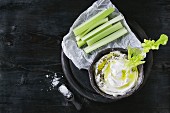 Fresh celery on paper with yogurt and olive oil dip in ceramic bowl, served with sea salt on wood round chopping board