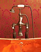 Edge of shiny copper bathtub and vintage tap fittings against red brocade wallpaper