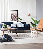 Kissen mit Denim-Bezug auf blauer Couch und auf Boden, Coffeetable, Stuhl mit Leder Auflage, Zimmerpflanze und Pendelleuchte im Wohnzimmer mit Retro Flair