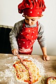 Kleiner Junge, als Koch gekleidet, belegt Pizza mit Würstchen