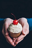 Hands holding a cupcake with peanut frosting and a cocktail cherry