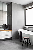 Freestanding bathtub in front of window and vanity in gray tiled bathroom