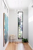 Heller Flur mit Armlehsntuhl vor schmalem Fenster, türkisfarbenes Bild an der Wand