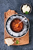 Bowl of red caviar on ice with spoon served with sliced bread, butter and herbs on terracotta board
