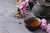 Black iron teapot and traditional ceramic cup of tea with blossom pink flowers cherry branch over gray texture background