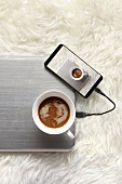 Tasse Kaffee auf Laptop daneben angeschlossenes Handy mit Foto von Kaffeetasse