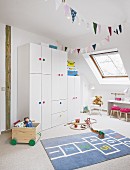 A white wardrobe with colourful handles and a hopscotch rug in a light and bright young girl's bedroom with slanted roof