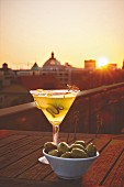 A glass of martini with olives at a rooftop bar as the sun sets