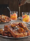 A plate of French Toast with sauteed bananas, butter and raisins drizzled with maple syrup on an old wooden table with fresh fruit cups in background