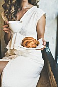 Young blond woman in white dress holding fresh croissant in plate and cup of cappuccino in cafe