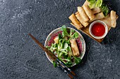 Fried spring rolls with red pepper sauces, served in traditional china plate with fresh green salad and wooden chopsticks over black texture background