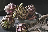 Uncooked whole organic wet purple artichokes in vintage bowl with textile sackcloth over dark wooden background