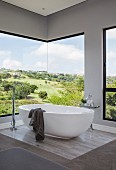 White free-standing bathtub in front of frameless, panoramic corner window with view of landscape