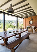 Long wooden dining table and bench in front of lounge area in interior with glass wall