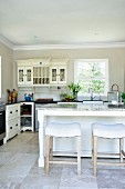 White stools and island counter in front of wall-mounted cabinet next to window in kitchen in French country-house style