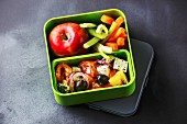 Griechischer Salat, Apfel und Gemüsesticks in Lunch-Box vor grauem Hintergrund