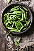 Young organic green pea pods and peas in terracotta tray over old dark wooden planks with sackcloth textile background